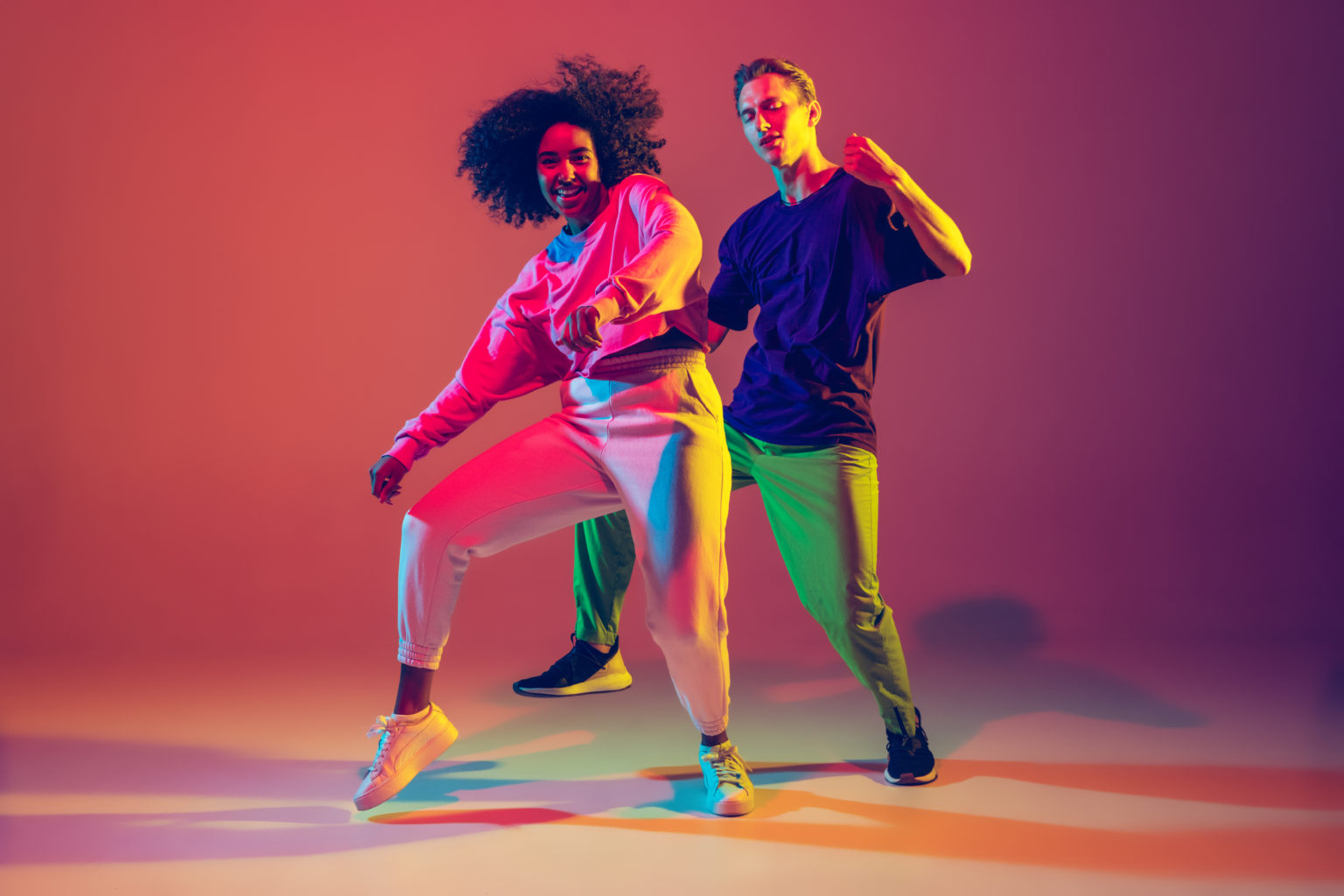 Dance time. Stylish men and woman dancing hip-hop in bright clothes on green background at dance hall in neon light. Youth culture, movement, style and fashion, action. Fashionable portrait.
