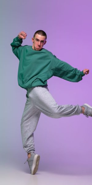 Beautiful sportive boy dancing hip-hop in stylish clothes on colorful gradient background at dance hall in neon light. Youth culture, movement, style and fashion, action. Fashionable bright portrait.